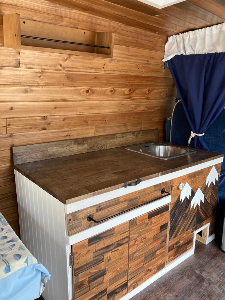 our kitchen for conversion van build. stained wood walls and cabinets. brown wood kitchen counter and sink. 