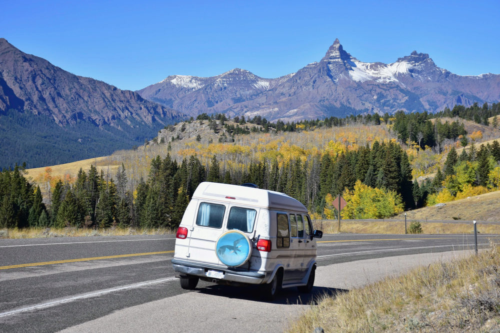 White van partially on the black asphalt roadway with bright green trees and snow capped mountains in the background. 