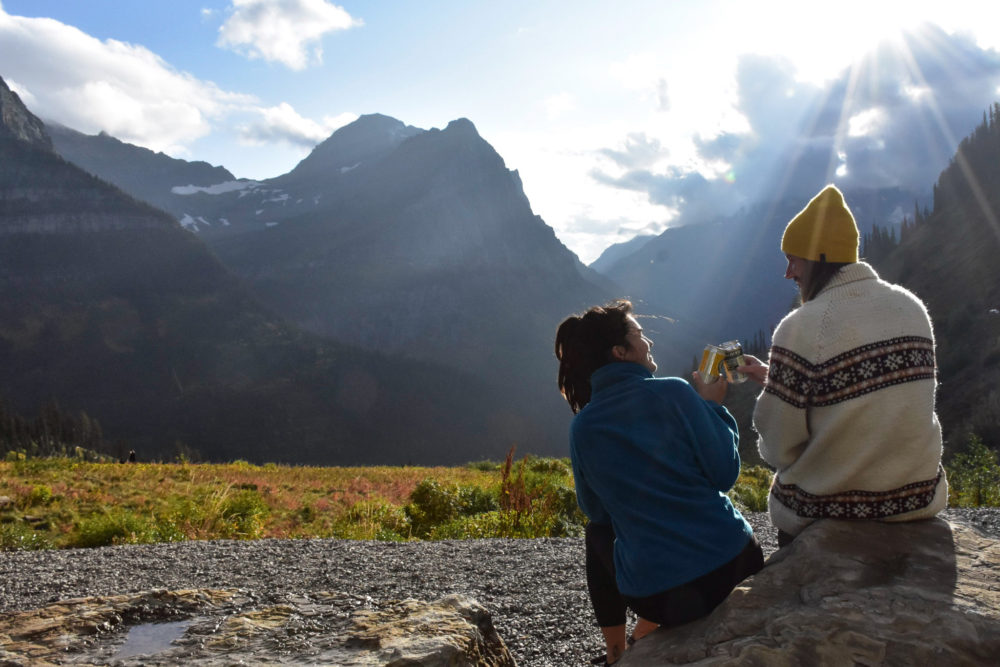 couple sitting on a cliffside cheersing beer cans and smiling. Van life with your partner