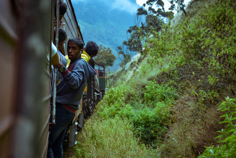Men hanging off the side of the train by bright green grass. 