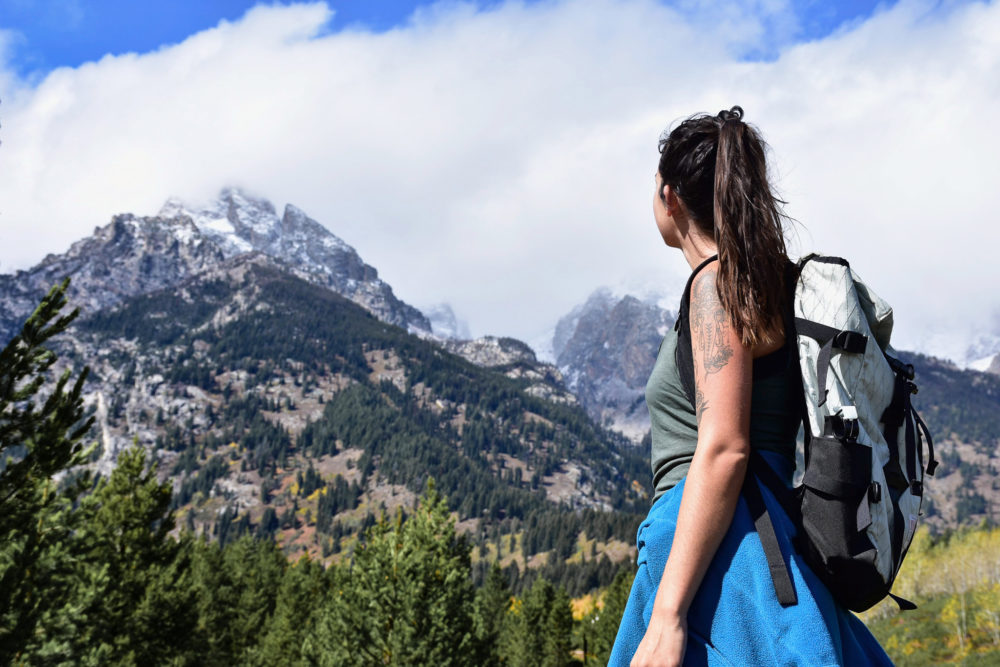 woman hiker looking up a snow-capped peaks of mountains and green trees around. 