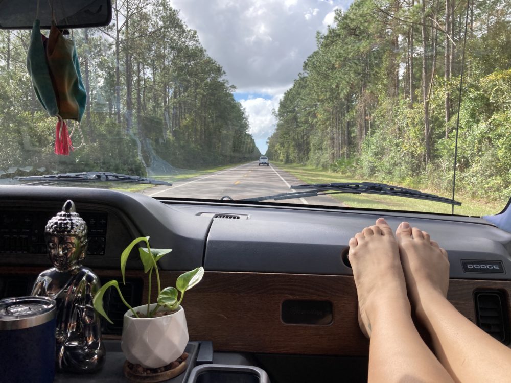 feet on the dashboard of a van with a plant and buddha statue visible inside and green jungle outside near the road. 