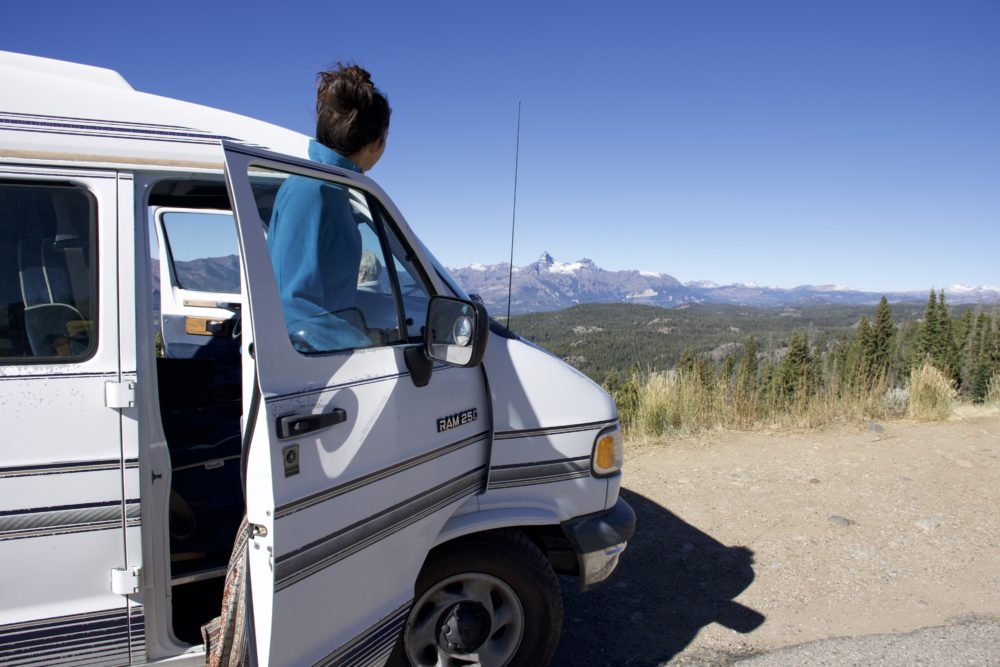 woman standing in doorway of white van looking at mountains and trees in the distance. benefits of van life 