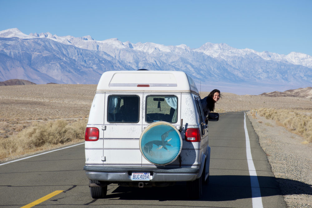 White van with a girl hanging out the window while driving down the road. Mountains in the background. Benefits of van life. 