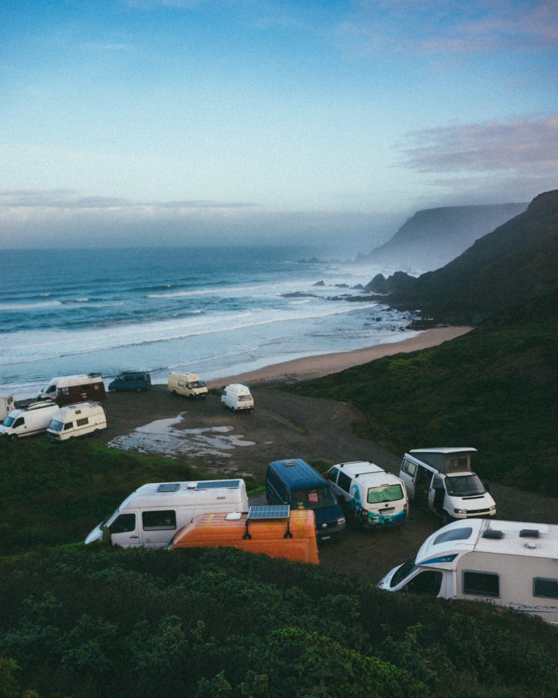 an orange van with solar panels, several white vans and a dark blue van parked on the beach with lots of green bushes surrounding it. reasons not to buy a sprinter for van life 