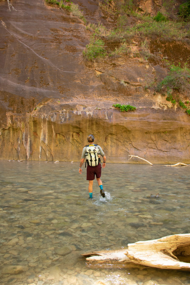 man walking through blue water river toward rock wall. 