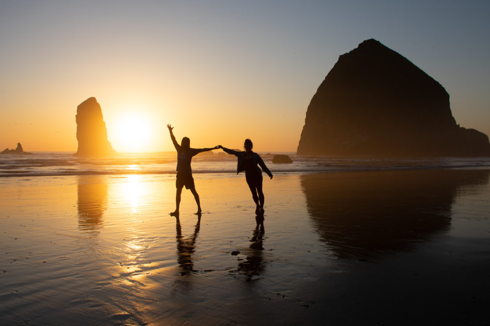 a couple posing with arms outstretched on a sandy beach at sunset with golden light and rock behind them. 