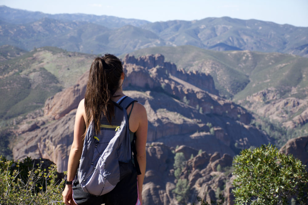 Woman with long brown hair and a backpack looking at a jagged cliff landscape below her. 