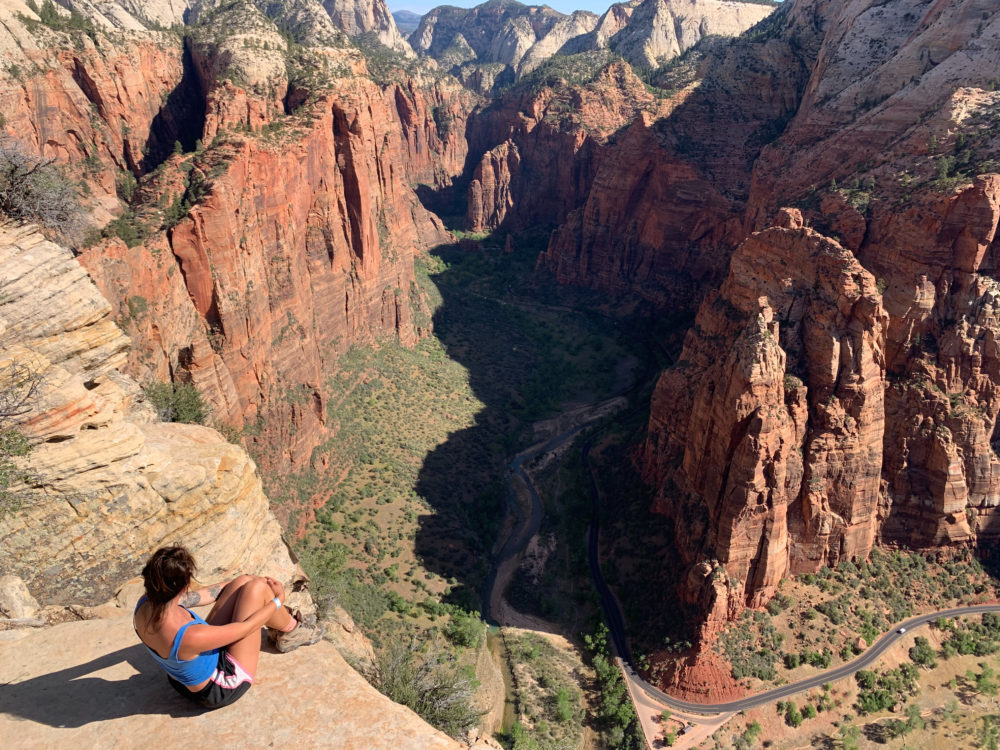 Girl sitting on the edge of a cliff overlooking green valley and red rocky mountains. 
