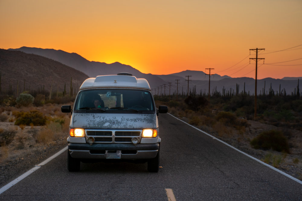 van driving on a road in desert with sun setting behind it