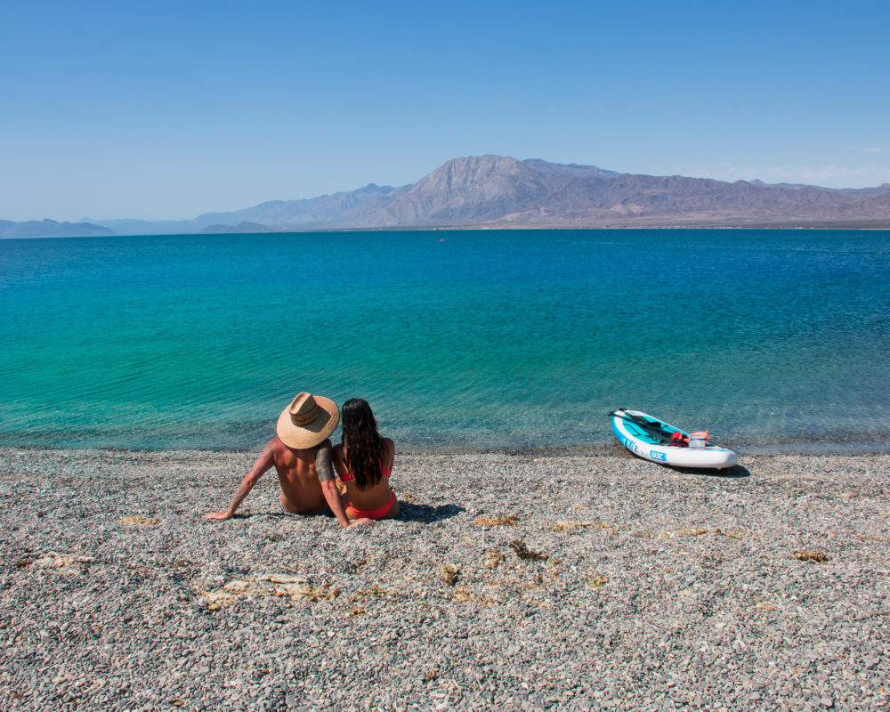 couple sitting on a pebble beach with bright blue water and a paddleboard 