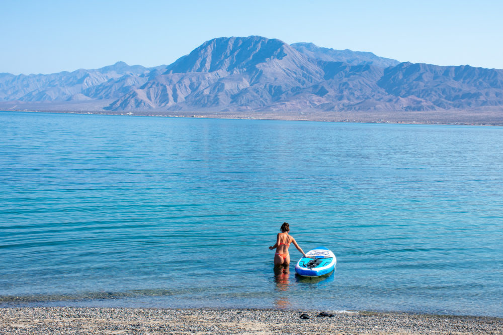 Woman with a paddleboard in the bright blue water with purpled hued mountains in the background. 