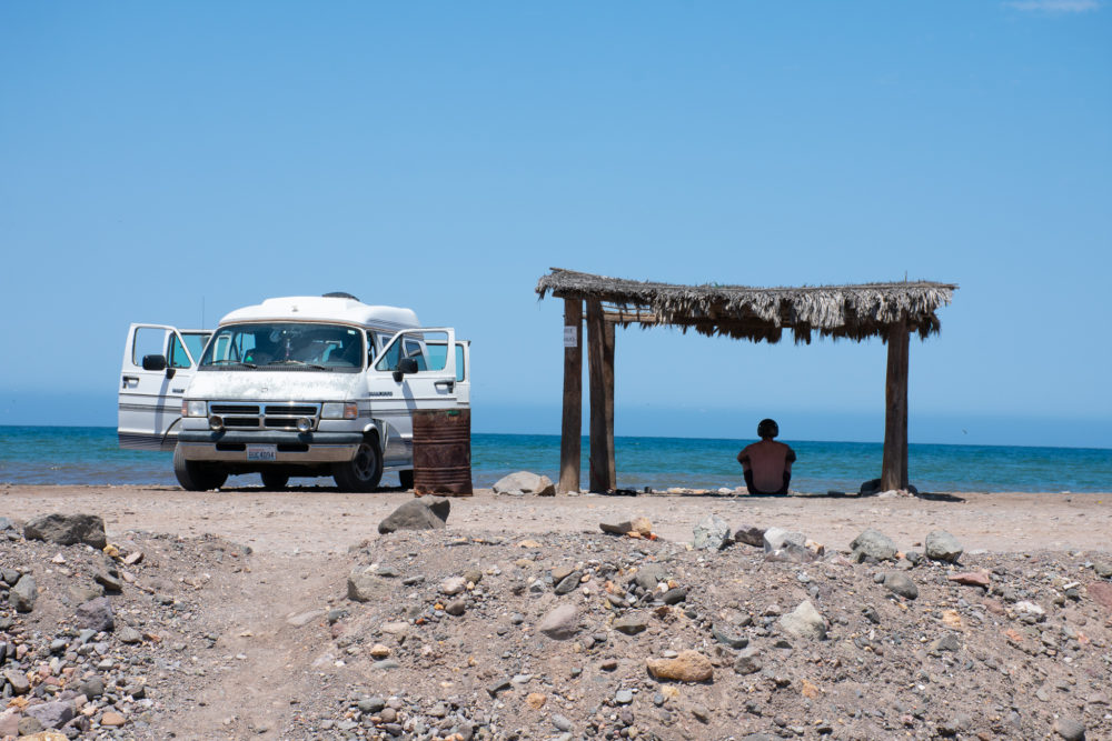 van parked on the rocky beach with a man under a thatched roof in the shade. 