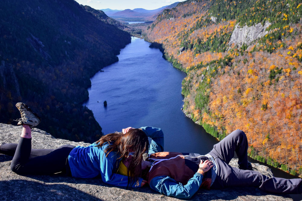 A scenic vista of a fall hike in the Adirondack mountains. A couple posing on a cliff overlooking a lake and fall colors below. 