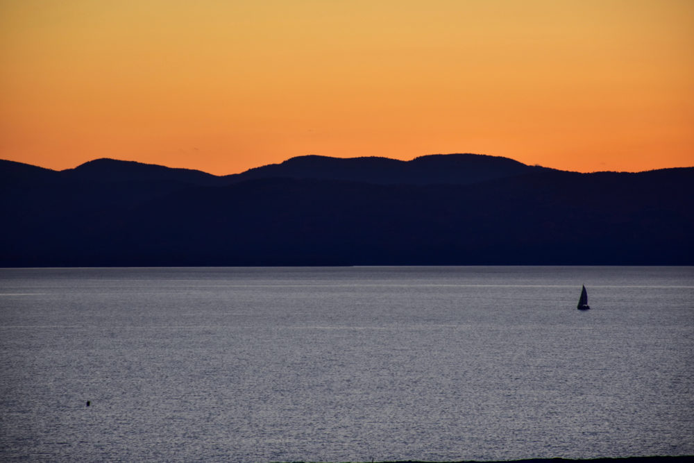 Lake Champlain at sunset. A small sail boat floats on the massive lake with dark hills and an orange sky in the background. 