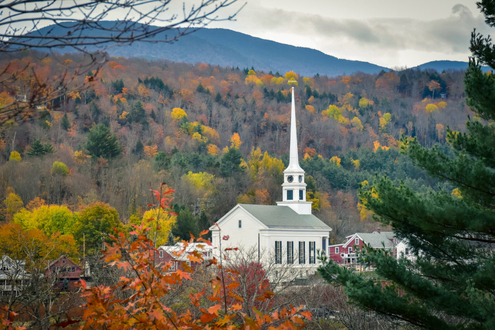 The town of Stowe, Vermont from afar. A massive white church with a thin steeple pokes out of the fall colors of the surrounding forest. 
