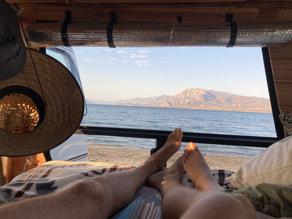 Couple of legs laying in a campervan bed looking out the window at a beautiful beach scenery. 