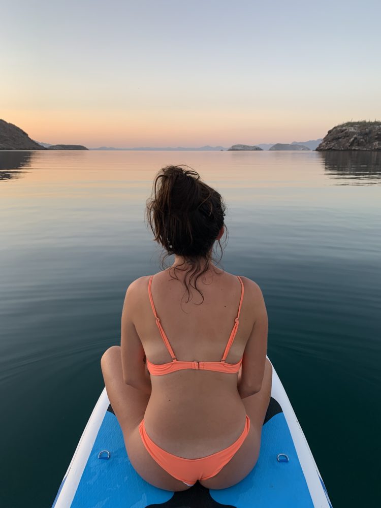woman sitting on a still ocean on a paddleboard with the colorful sunrise in the background. 