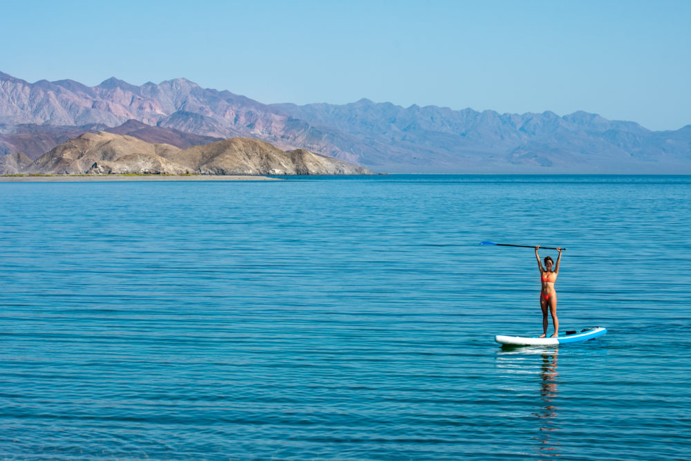 woman in an orange bikini standing triumphantly on a paddleboard in the ocean off Baja Mexico. Things we love about our campervan