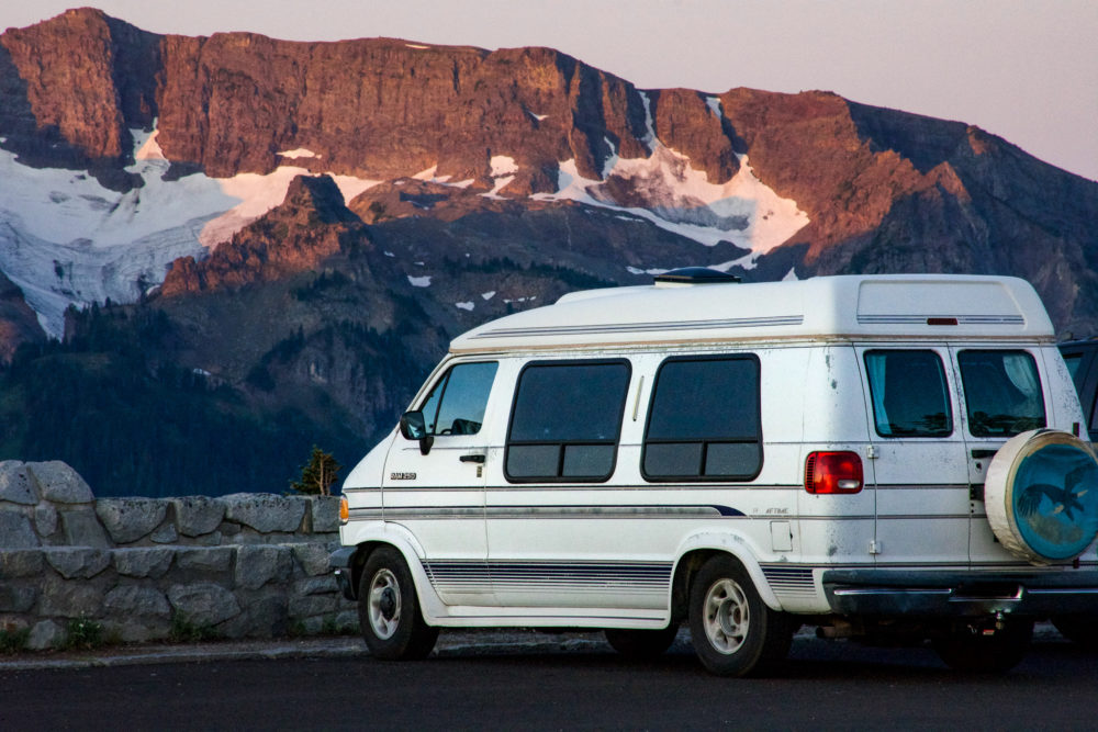 A white van used as a camper is parked in a parking lot looking at the mountains in Rainier National Park. 