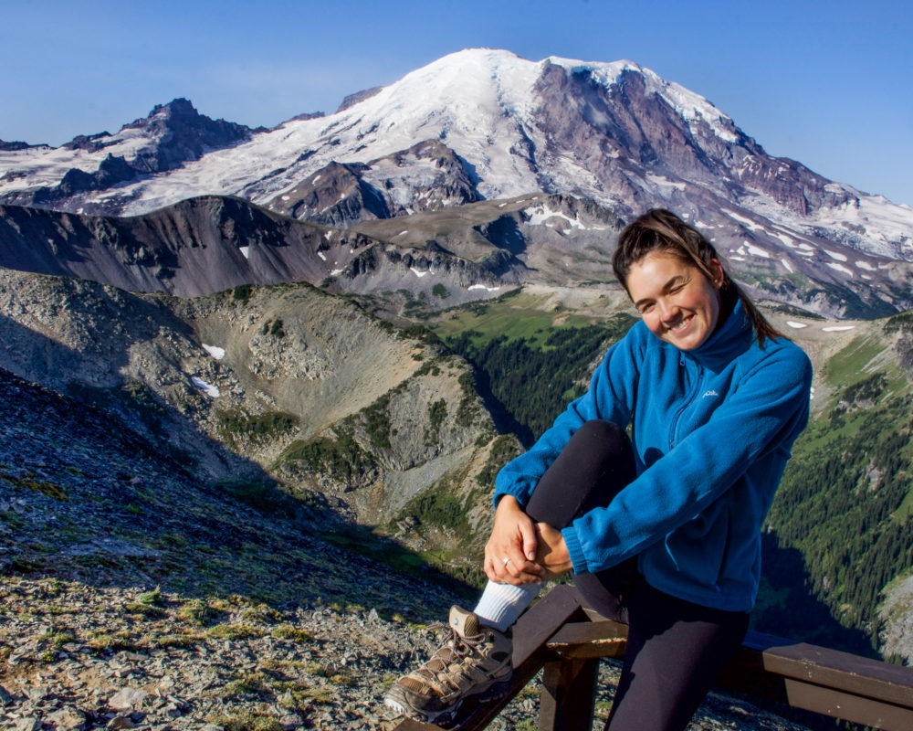 The view from Fremont Fire Lookout Trail in Mount Rainier National Park. Mount Rainier is looming in the background and a woman poses on the railing of the lookout. One of the best hikes for a weekend in Mount Rainier National Park 