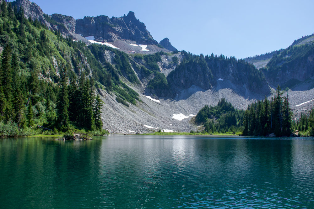 The landscape of Snow Lake, one of the best hikes for a weekend in Rainier National Park. Icy blue water sits surrounded by snow capped peaks and green trees. 