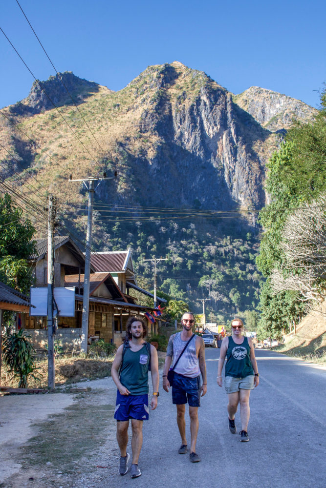 three people walking down a dusty street with mountains in the background 