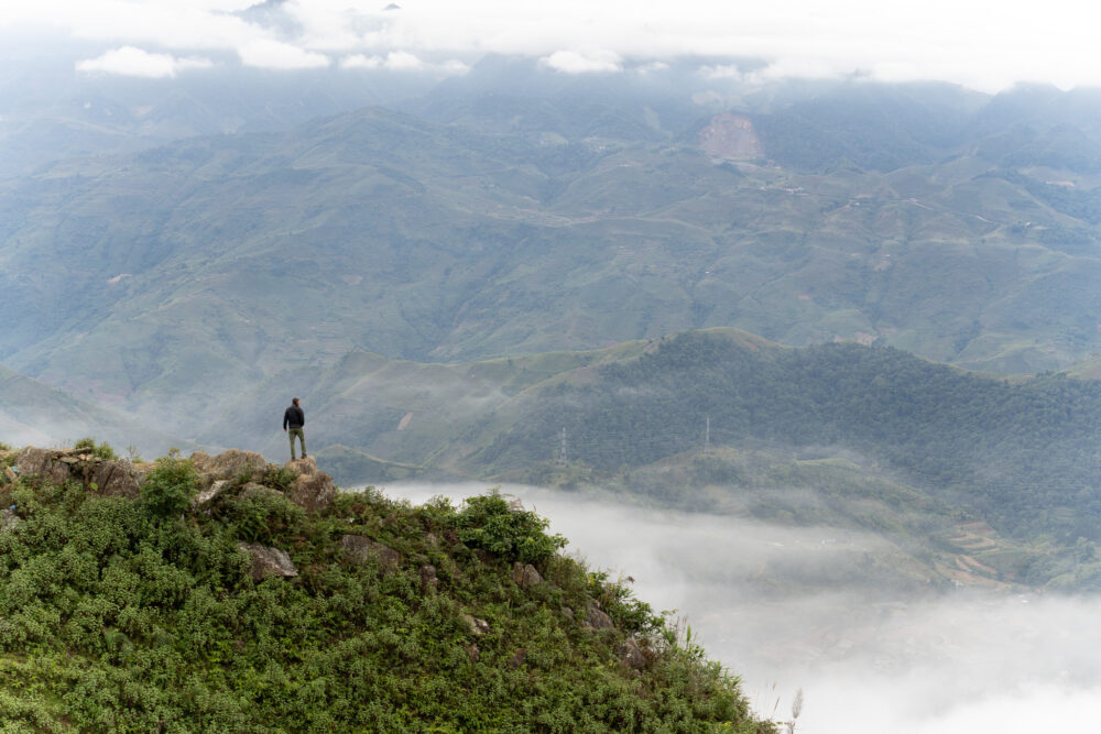 A man standing on a cliff with panoramic views of the green mountains and clouds behind him. 