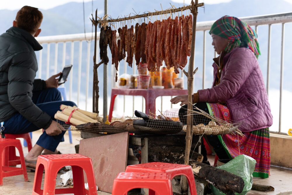 Hmong women in traditional dress serving food with dried meat by a fire. 