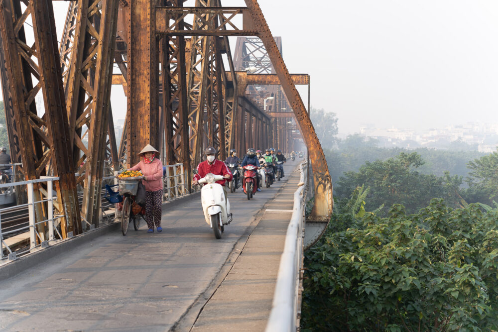 Motorbikes driving on a bridge above the banana trees passing a woman pushing her bike filled with oranges. 