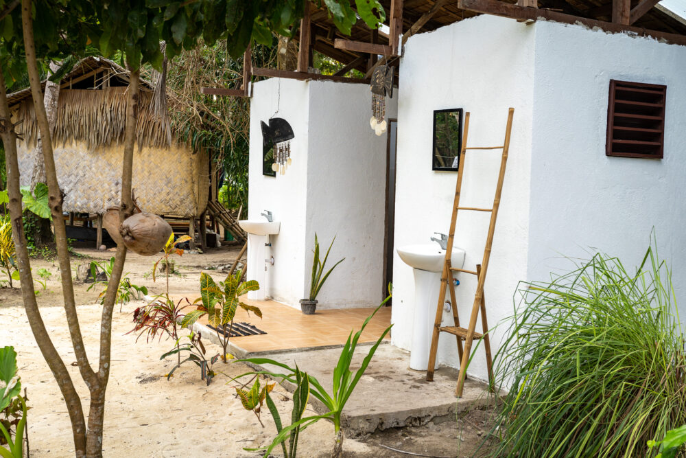 White outdoor bathroom at Isla Experience area with trees an a ladder leaning on it. 