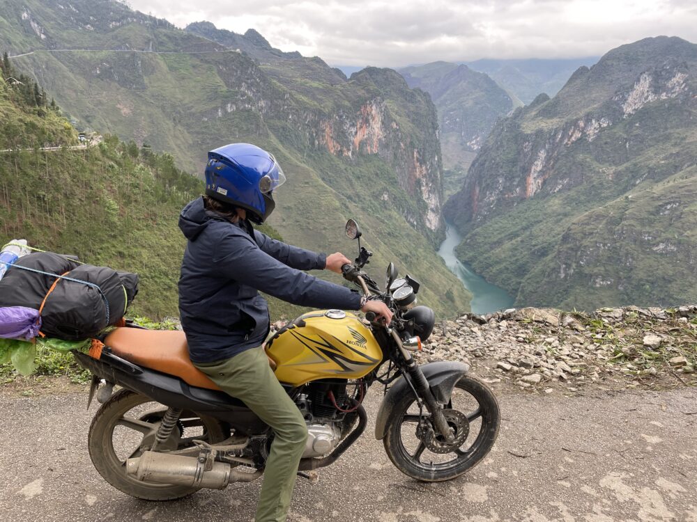 Man on a gold motorbike overlooking a canyon with red rocks, green hills, and a blue river inside. 