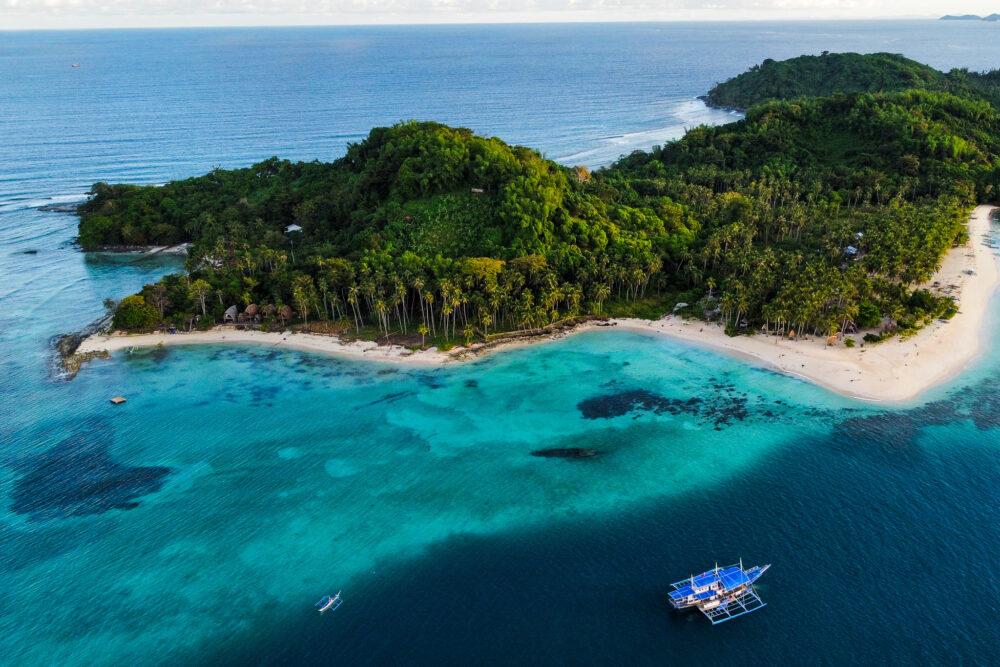 Aerial view of a lush green island with white sand beaches, blue water, and a small boat approaching. 