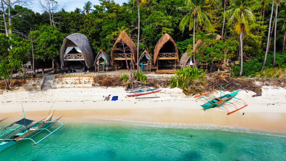 An aerial view of a beach with boats parked in it and traditional huts behind it at the Isla Experience