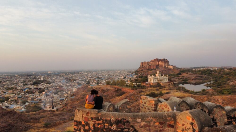 A couple sitting on an ancient wall at sunrise looking at the view of a marble building and a massive fort in the distance. 