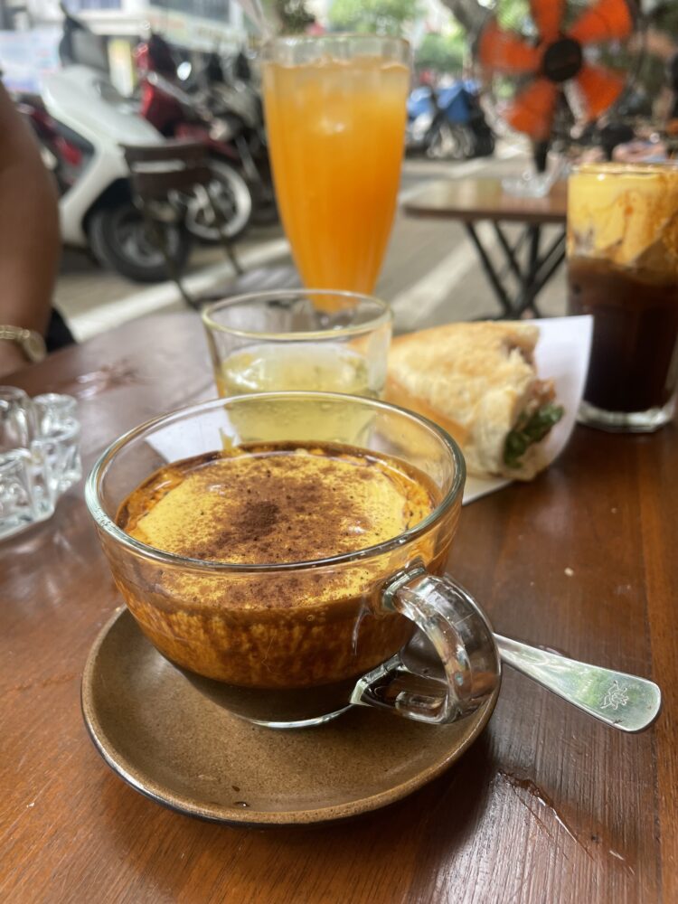 a frothy egg coffee on a table with juice and a sandwich behind it. 
