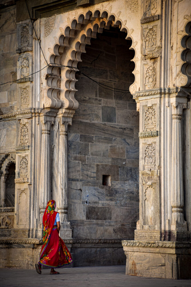 Woman in a red sari walking beneath an intricate carved gate. 