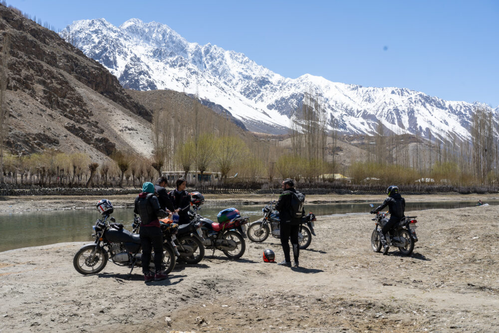 A group of motorbikes sit by a river with a snowy mountain behind them. 