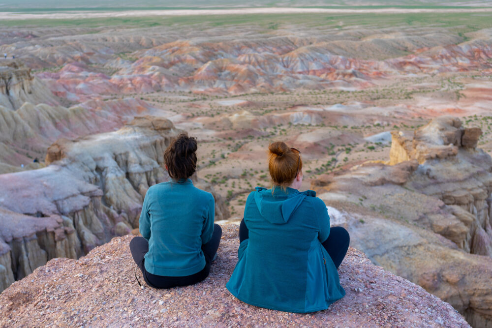 two girls in blue jackets sitting on the edge of a desert land formation in white stupa 
