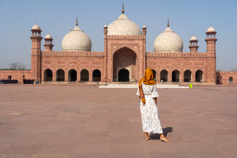 woman in an orange head-scarf wlaking in front of the Badshahi mosque 