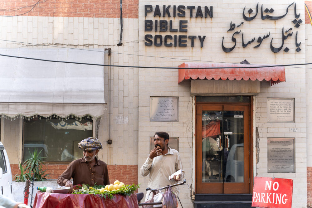 Two men stand on the street in front of the pakistani bible society sign 