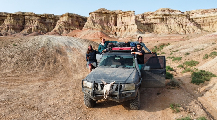 four people hanging out the doors of a 4x4 vehicle with desert cliffs behind him