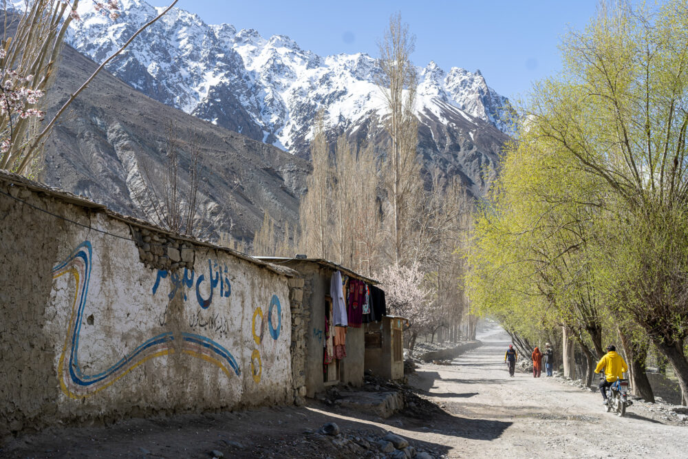 a small town in northern pakistan with people walking on a dirt road and huge high mountains 