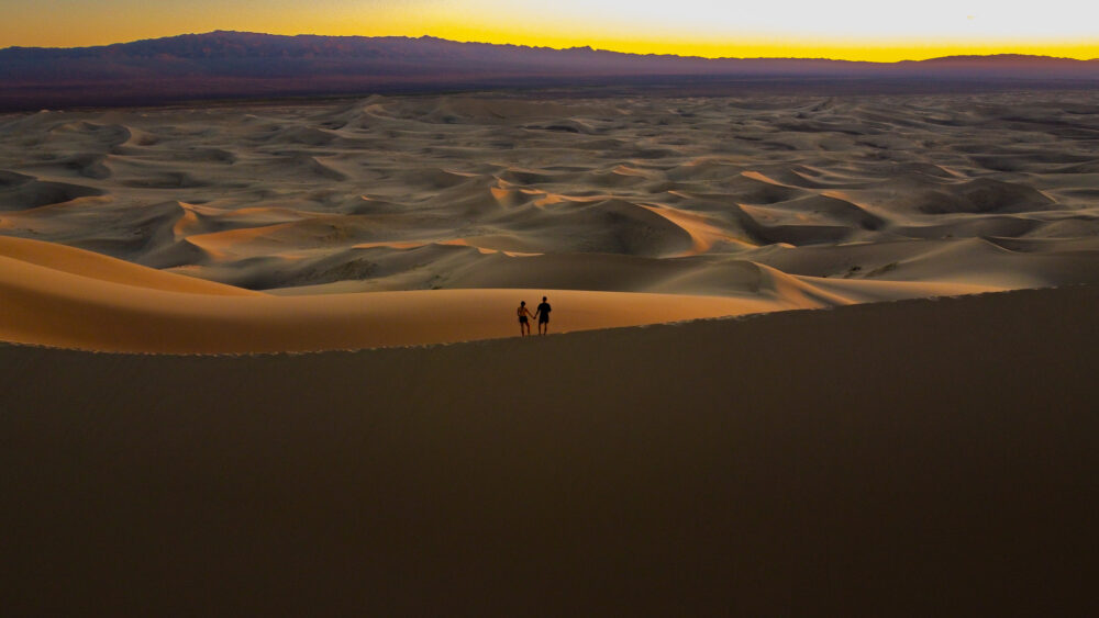 two people standing on top of a huge sand dune at sunset