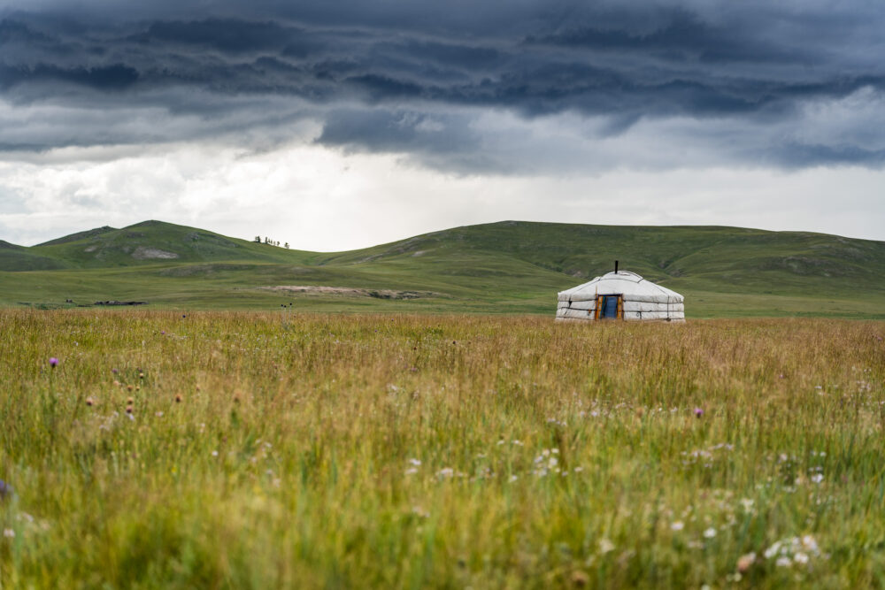 one single ger sitting in a wide open field with clouds above it