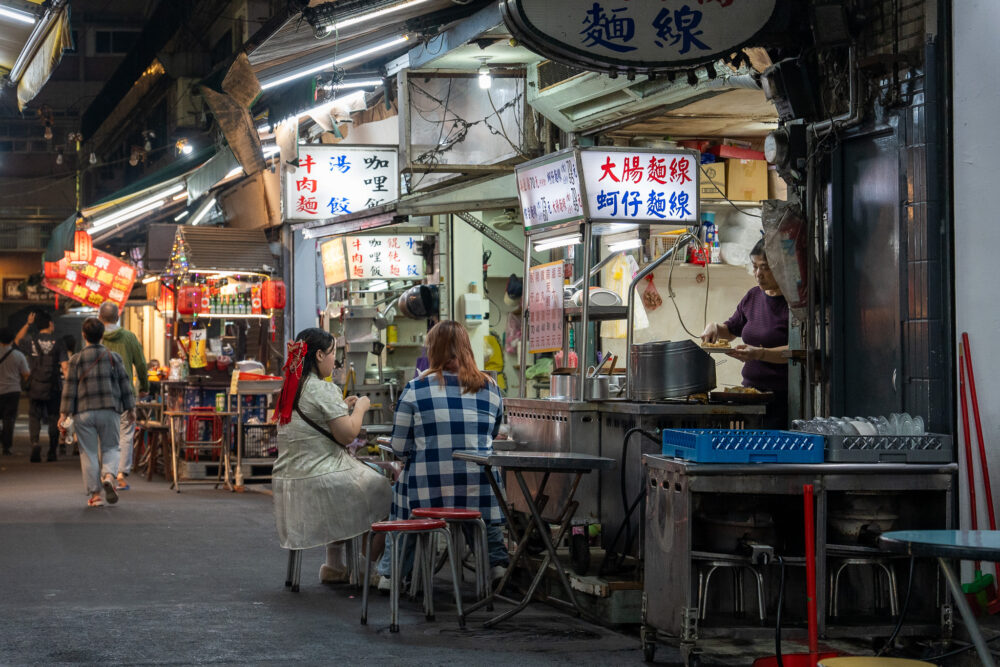 two women sit in front of a food stall at night eating in Taiwan
