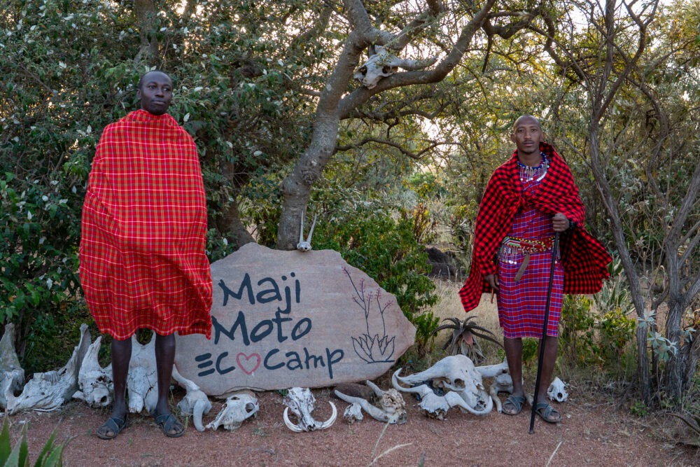 two Maasai men pose by the entrance sign to Maji Moto Eco Camp 